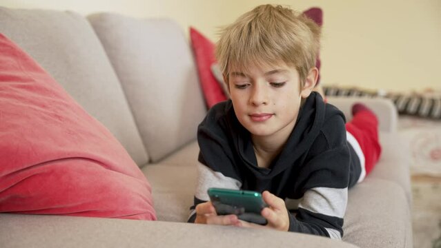 Boy lying on the sofa watching video or photo content in social network, scrolling by smart phone, smiling child enjoys mobile app at home. Online education, communicating distantly, playing games
