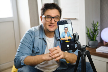 Hand free. Middle-aged male sitting at home before smartphone fixed on tripod. Video blogger recording content to channel. Young man making video stream