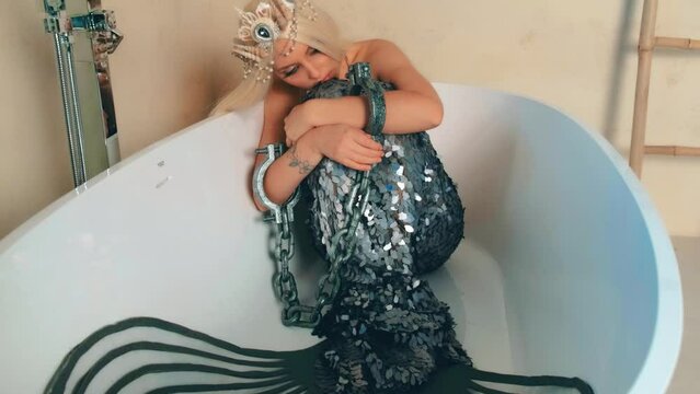 Closeup portrait art fairytale woman mermaid. Girl nymph captive sits in bathroom. On hands shackles, chain. Lady mistress seas hugs tail. Blonde long hair. Silver creative carnival costume, fishtail