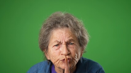 Closeup portrait of angry upset frustrated angry elderly senior old woman saying shh, quiet, with wrinkled skin and grey hair on green screen background.