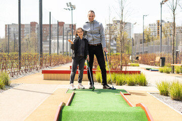 Fototapeta na wymiar Sharing with golf experience. Cheerful young man teaching his daughter to play mini golf at the day time. Concept of friendly family