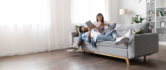 Relaxed indian spouses resting on sofa at home, using gadgets
