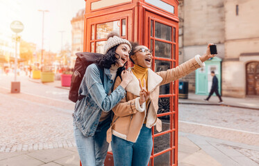 two friend, girlfriend and women using a mobile phone, camera and taking selfie against a red phonebox in the city of England.Travel Lifestyle concept