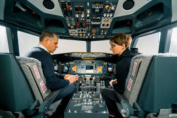 An experienced pilot instructs a young student and shows a small model airplane in the cockpit of an aero simulator