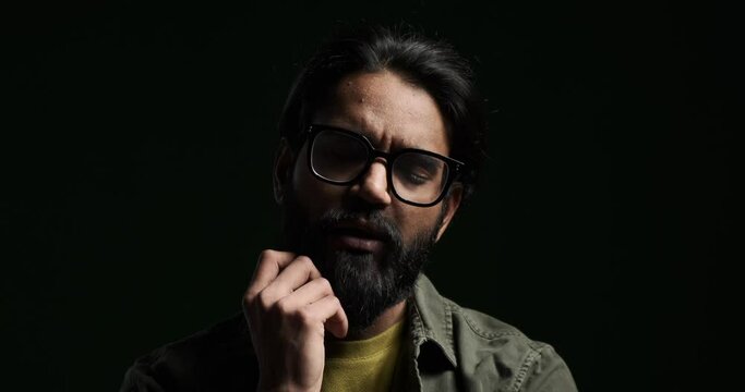 Closeup portrait of serious thoughtful young man touching beard and looking at camera isolated against black background