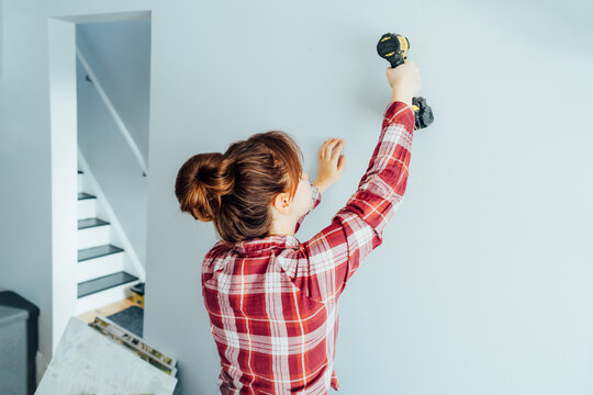 Back view young brunette woman screwing screw into wall with electrical screwdriver. Girl wants to put a picture on the wall. Housekeeping work. Doing repairs herself, DIY, gender equality concept.