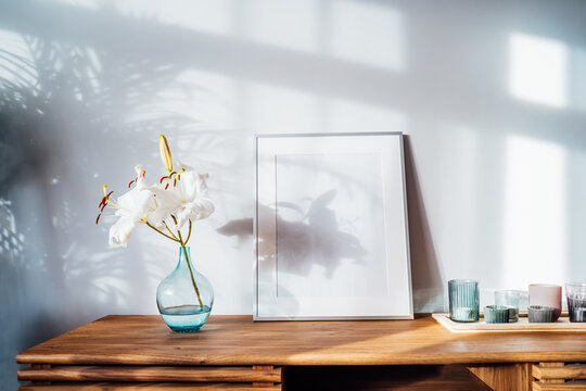 Modern minimalist Scandinavian style interior with white poster mockup, candles and white lily flowers in vase on a wooden console under sunlight and home plants shadows on gray wall. Selective focus.