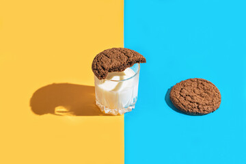 Creative composition made of brown american cookies and a glass of milk on blue and yellow background. Minimal style. Bakery and pastry concept. Idea for breakfast.