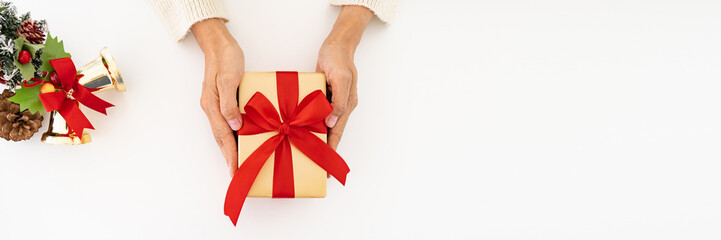 Woman hands in white sweater, on white table background with christmas tree and bell, giving  Christmas holiday golden gift  box with red ribbon. -  top view with copy space Banner