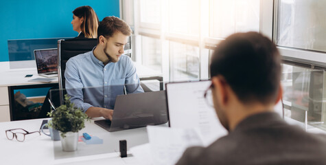 Concentrated on work. Concentrated young beard man working on laptop while sitting at his working place in office