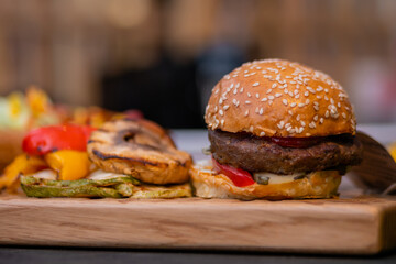 Fresh large burger and grilled vegetables: zucchini, bell pepper on wooden cutting board on counter for sale at summer local food market - close up. Outdoor cooking, gastronomy and street food concept