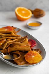 Traditional French Crêpes suzette pancakes with orange sauce on a serving dish on a light grey tale background decorated with citruses and bloody oranges.