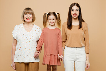 Obraz premium Happy smiling cheerful women wear casual clothes with child kid girl 6-7 years old. Granny mother daughter holding hands looking camera isolated on plain beige background. Family parent day concept.