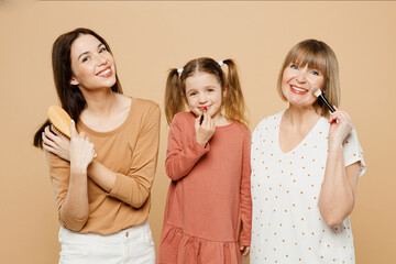 Happy smiling fun women wear casual clothes with child kid girl 6-7 years old. Granny mother daughter do make up comb hair use lipstick isolated on plain beige background. Family parent day concept.