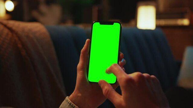 Indoors hands man holding and using a mobile phone with a vertical green screen lying on a sofa. Social media. Mock up