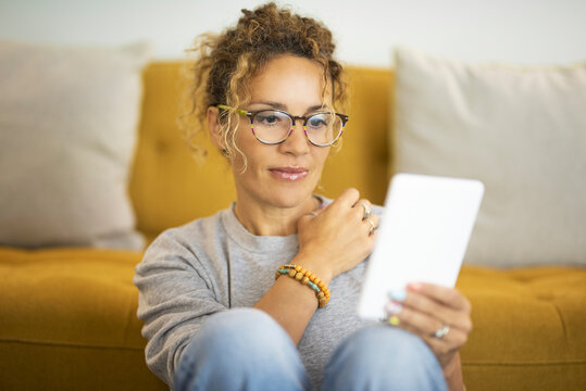 Front view of woman using tablet at home in relaxation lifestyle activity. Cute female people alone sitting against yellow sofa in indoor leisure technology connection activity. Modern lady eyewear