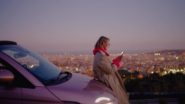 Smiling female influencer surfing social media on mobile phone while sitting on car at evening with scenery flickering city view.Cheerful young woman using smartphone application while browsing online