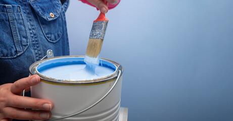 woman wearing overalls taking out the brush from the jar of blue paint next to a blue wall