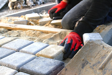 Paver master in red gloves. Man lays paving stones in layers. Garden brick pathway paving by professional worker. Hands of worker installing concrete paver blocks by rubber hammer. Out of focus - obrazy, fototapety, plakaty