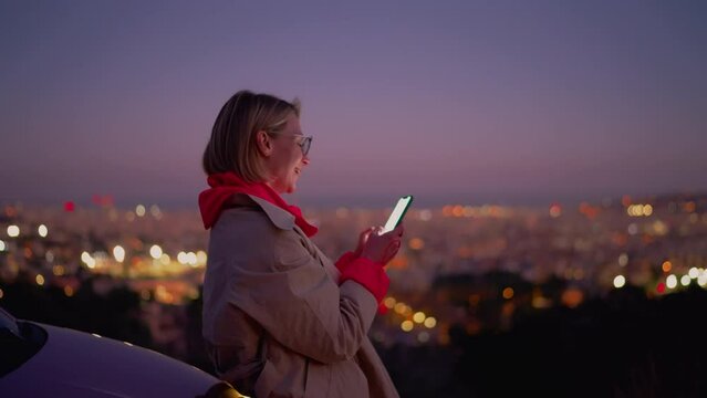 Relaxed content creator stands at panoramic night city view,working on latest blogging post using trusty mobile phone application. Young smiling female using smartphone for communicate with her family