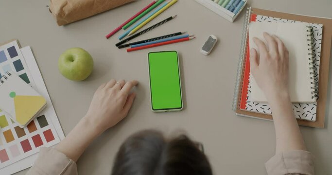 Top view of desk with stationery and chroma key green screen smartphone and female artist sitting at table. Workplace and design application concept.