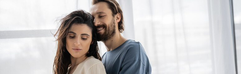 Positive bearded man standing near brunette girlfriend at home, banner.
