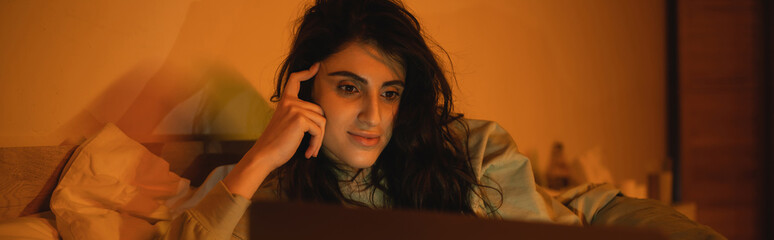 Fototapeta premium Brunette woman watching film on blurred laptop in bedroom at night, banner.