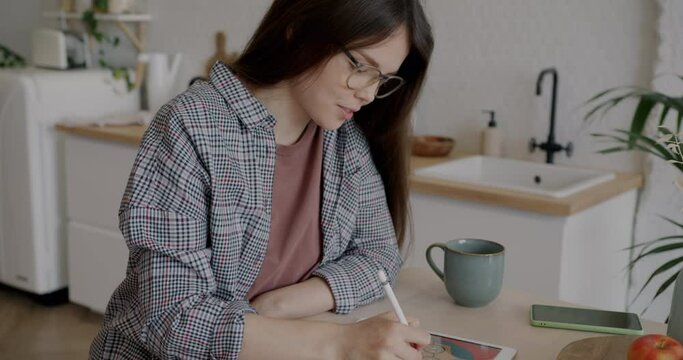 Smiling young woman illustrator drawing with digital graphics tablet sitting at table in kitchen. Creative occupation and gadgets concept.
