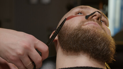 Barber is cutting beard of man using scissors in professional barbershop.