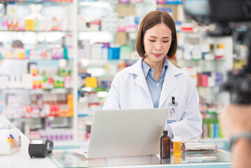 Professional pharmacist asian woman smiling at counter using laptop computer for check medicine inventory in drug store, Medical service online, Telemedicine to giving medical information to patient.