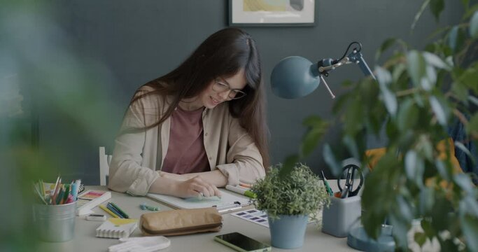 Ambitious young designer drawing and smiling enjoying work in creative art studio. Cheerful woman sitting at desk in cozy office and using crayons and paper.