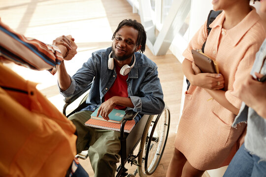 African American student with disability sitting on wheelchair and greeting his classmates during break at college