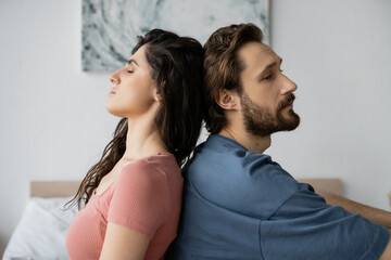 Upset bearded man sitting back to back with brunette girlfriend in bedroom.