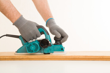 Adult man hands using electric jointer and shaving old wooden plank for furniture or floor on white table at light gray wall background. Closeup. Preparing material for repair work of home. Side view.
