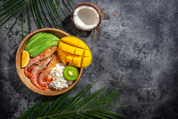 Poke bowl with avocado, shrimp, rice, mango, kiwi and coconut on a dark background, healthy and balanced food. banner, menu recipe place for text, top view.