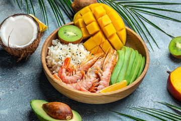 tropical bowl with avocado, prawns, rice, mango, kiwi and coconut, tropical food on the sea beach on light background with tropical leaves. Healthy food, clean eating, Buddha bowl, top view