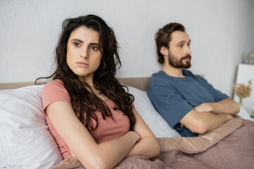 Nervous woman crossing arms while sitting near blurred boyfriend on bed.