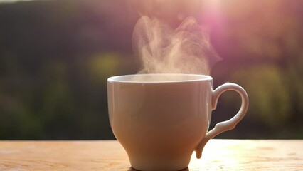 A cup of hot coffee on the background of the morning sunrise. The steam rises up and is illuminated by the sun's rays. Warm sunset colors and cups of tea.