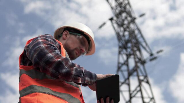 A power engineer inspects a high-voltage power line and records electrical sensor data on a tablet. Energy crisis in the country.