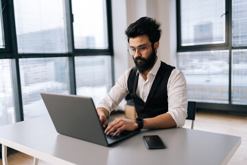 Bearded Indian business man in formalwear working typing on laptop sitting at desk in light coworking office on background of window. Bearded freelancer male in eyeglasses looking at computer screen.