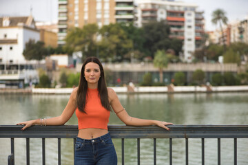 Young and beautiful woman with straight brown hair, orange top and jeans, leaning on a railing looking at the camera with a calm gesture. Concept fashion, beauty, trend, peace, relax, millennial.