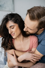 Bearded man hugging smiling brunette girlfriend in bedroom.