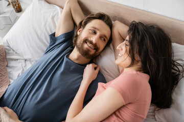 Top view of smiling brunette woman looking at bearded boyfriend on bed in morning.