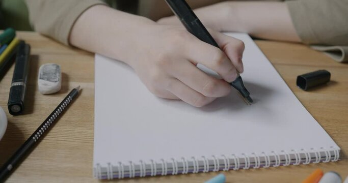 Close-up of female hand drawing with marker on paper while woman enjoying hobby indoors at table. Art and creative activity concept.