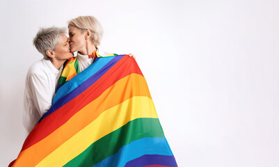 A couple of short-haired women between 50 and 60 years old, very much in love and embracing each other, covered by the gay or LGBTQ+ pride flag. Studio shot on a white background
