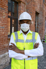 Serious man in reflective uniform and protective mask crossing arms near unfinished building. Portrait of african american man confidently looking at camera outdoors.