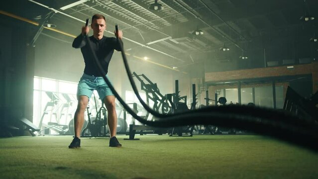 Athletic handsome young man doing slams on battle rope in fully equipped gym. Man wears black long sleeve compression shirt and sneakers, blue shorts. High quality 4k footage