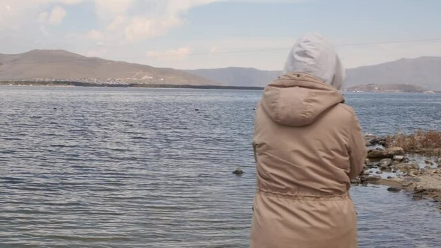 A young woman in a sand-colored jacket and wearing a hood walks along the seashore with high mountains on the shore on a sunny but windy day, looking behind