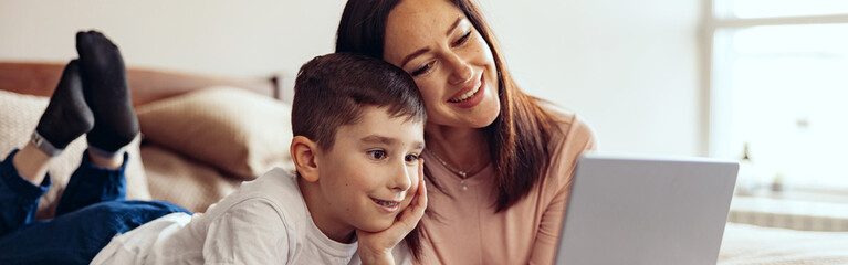 Pretty Caucasian mother and small teen son lying on bed and videochatting on laptop.