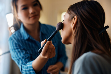 Close up of Asian makeup artist applying blush on young woman's cheek.
